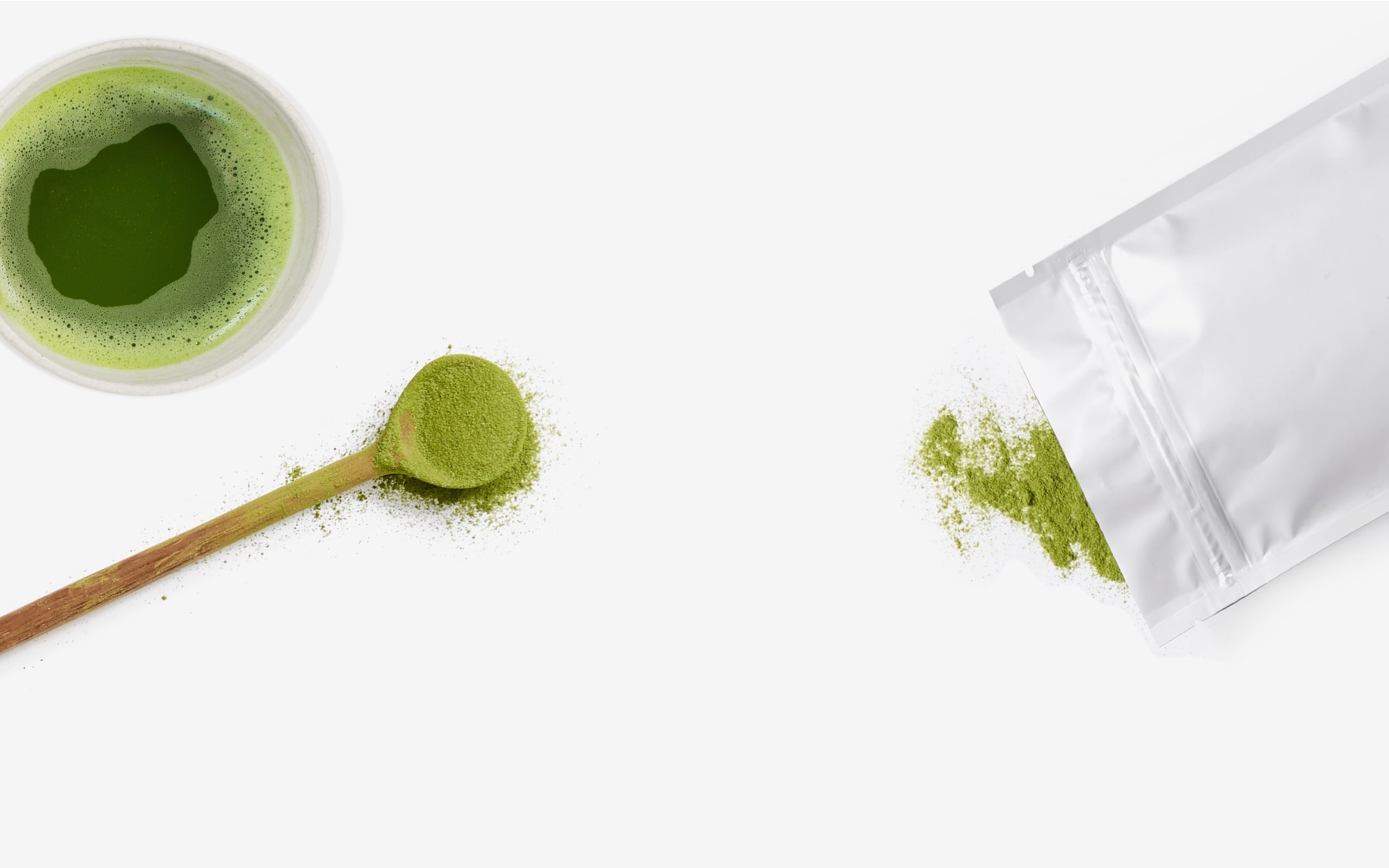 A cup filled with matcha next to a wooden spoon and bag filled with matcha.