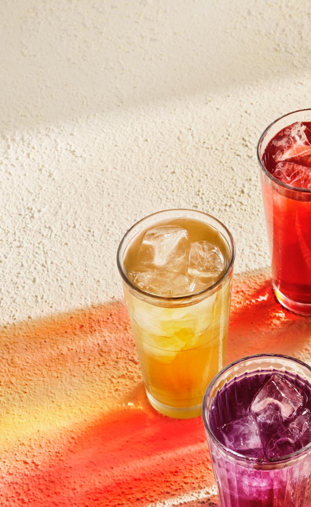 Seven glasses filled with various colourful iced teas emanating their colourful shadows.