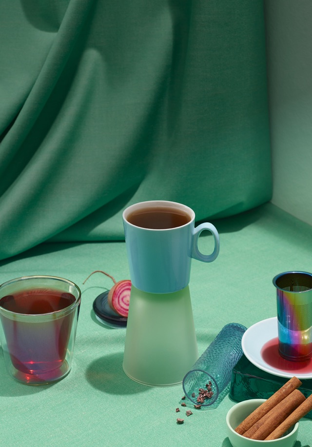 Des tasses variées remplies de thés sur une surface turquoise et entourées de bâtons de cannelle et d’un infuseur arc-en-ciel.
