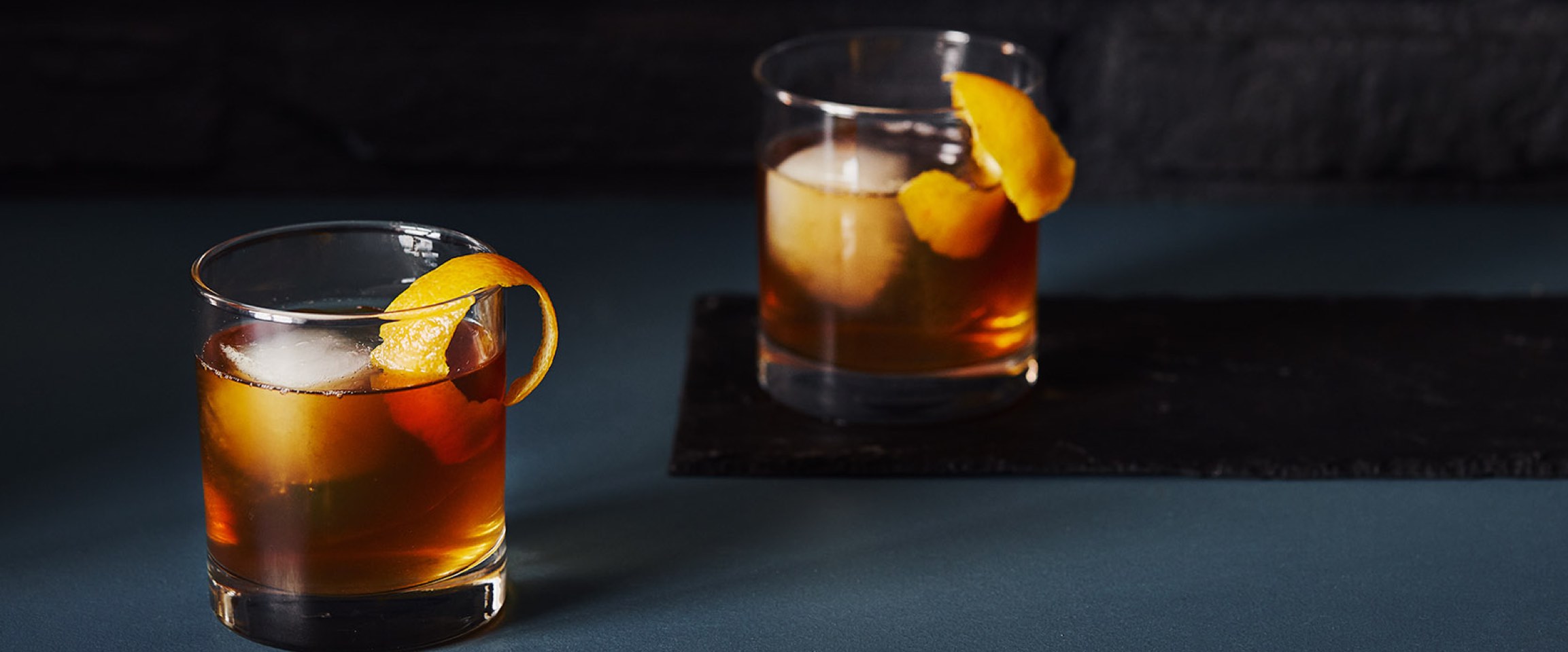 2 clear glasses with Manhattan cocktail, ice and orange peel.